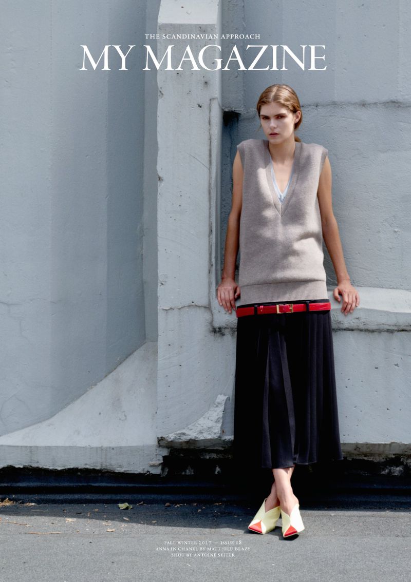 Fashion model leaning against a grey concrete wall wearing a sleeveless knit vest, red belt, black pleated skirt, and yellow mules.