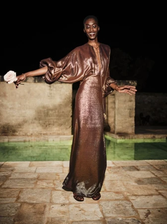 A Black woman in a copper gown holds a rose, photographed outdoors at night with a stone pool in the background, smiling gently.