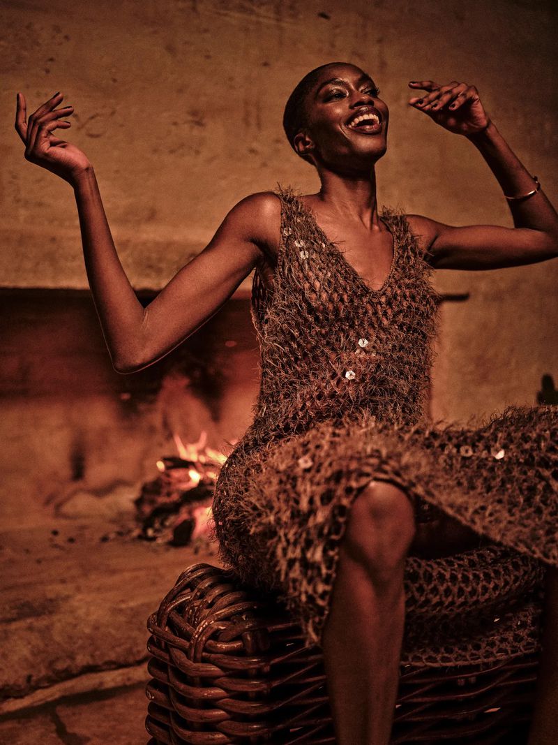 Smiling African-American woman in a brown, textured dress, seated on a woven basket-like stool in front of a stone fireplace indoors.