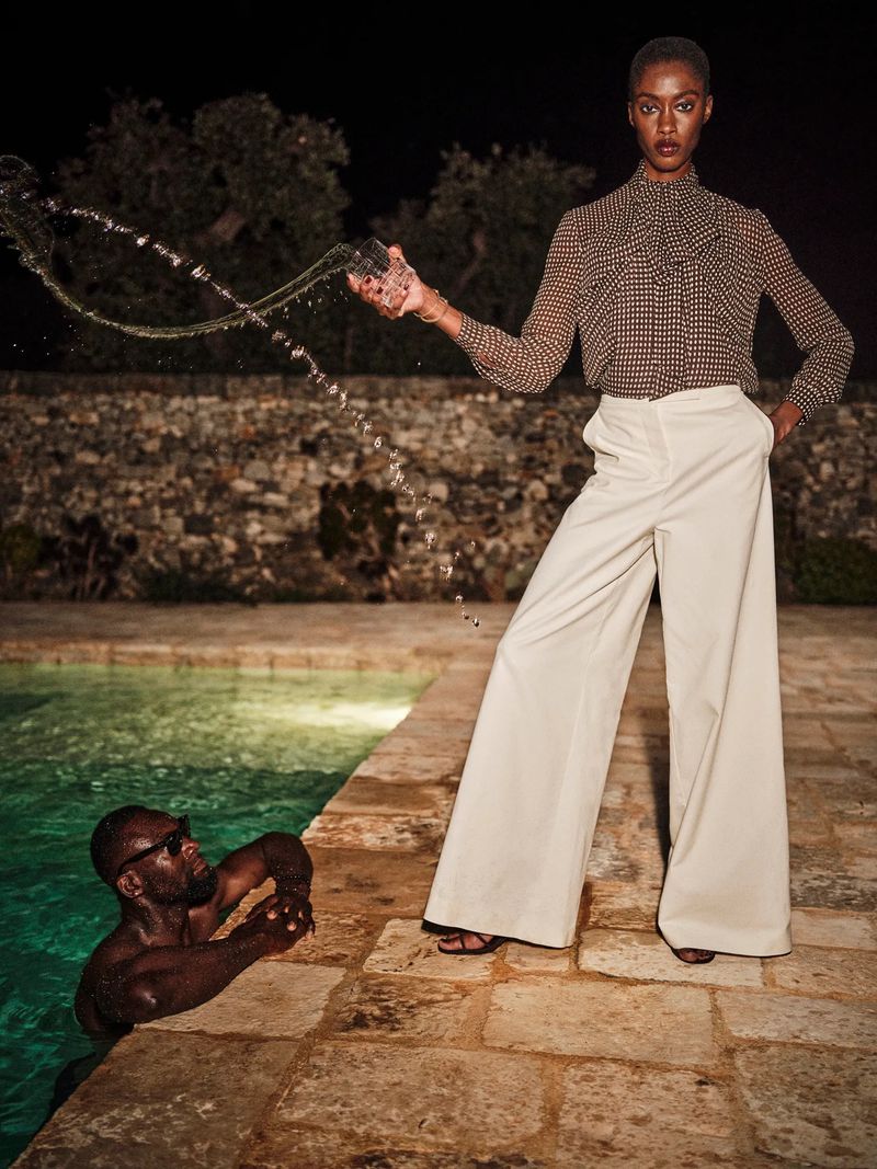 A tall African-American woman in cream pants and patterned blouse holds a glass, standing near a pool with an African-American man.