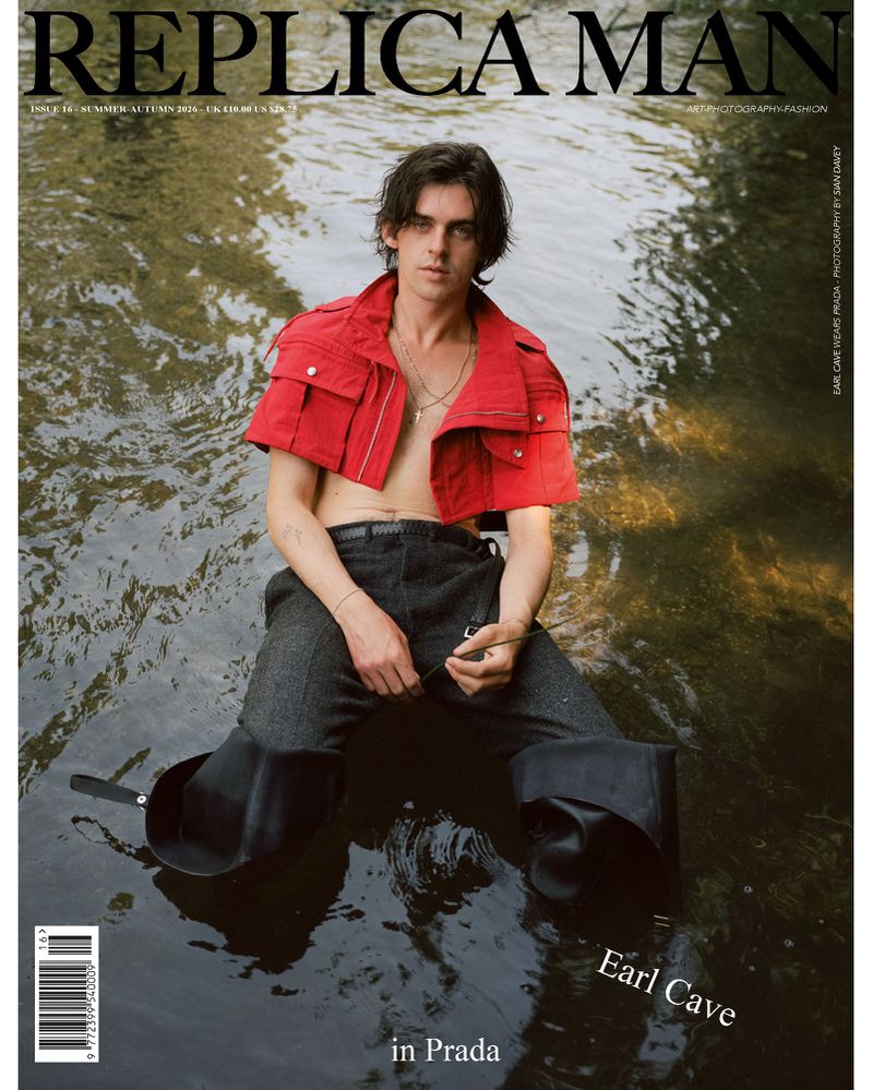 Earl Cave wearing a red cropped jacket and dark trousers seated partially submerged in natural river water for Replica Man Magazine.