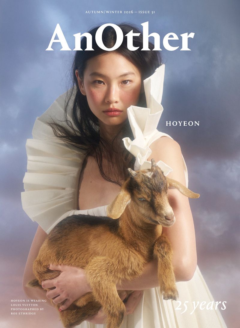 Hoyeon in a sculptural white pleated dress holding a brown goat kid against a cloudy backdrop on the cover of AnOther Magazine.
