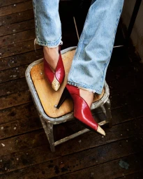 Close-up of red high heels with pointed toes resting on a wooden stool, wearing light wash blue jeans.