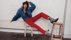 Model wearing an oversized denim jacket, red jeans, and animal-print boots seated across two wooden stools in a studio setting.