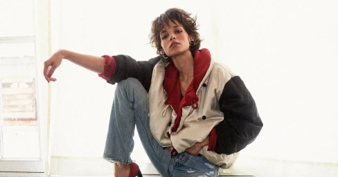 Model with short dark hair wearing an oversized colorblock jacket and blue jeans, seated against a bright window backdrop.
