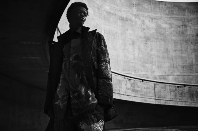 Black and white fashion shot of an African-American man in an overcoat with a patterned vest in front of a concrete wall.