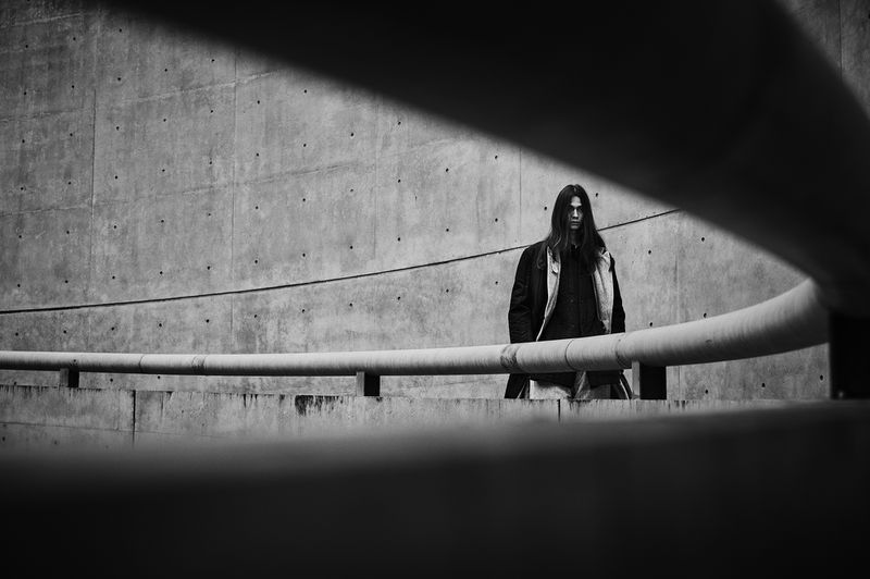 Monochrome portrait of a slender adult male with long dark hair wearing a coat, standing near a concrete wall and metal railing.