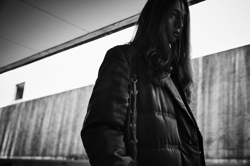 Black and white portrait of a long-haired person in a padded jacket with a chain detail, looking to the side with a concrete wall background.