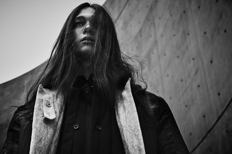 Monochrome portrait of a long-haired person wearing a black coat with fur accents against a textured wall, captured in low angle.