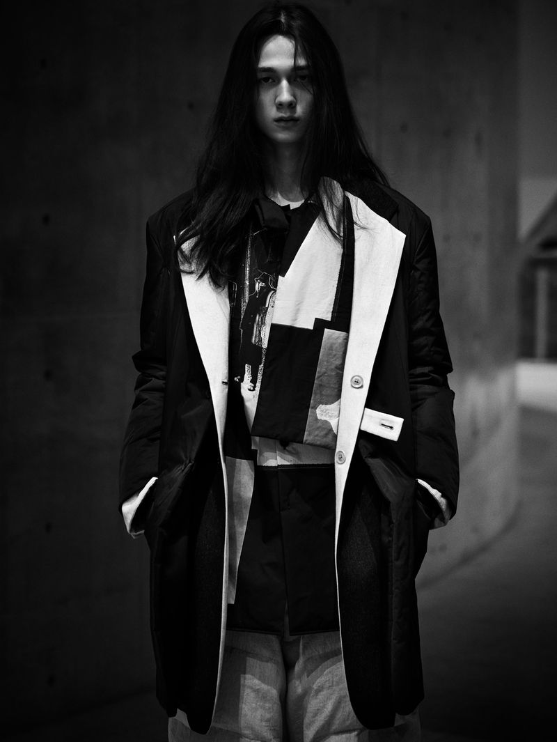 Monochrome portrait of a long-haired male wearing a black and white patterned top with blazer, standing indoors against a dark background.