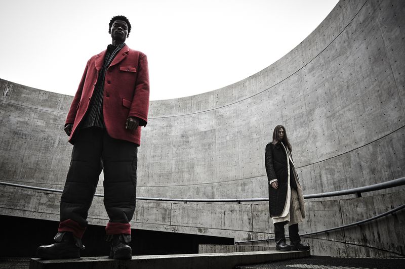 Two people stand in an urban outdoor setting with concrete architecture; one wearing a red jacket, the other a black coat, shot at eye level.