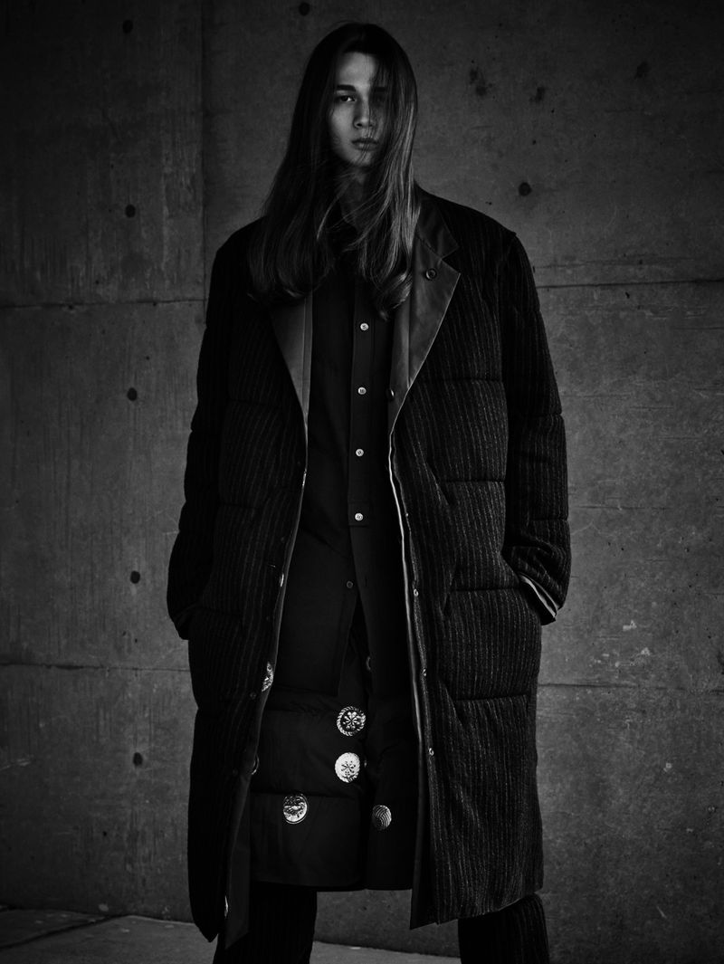 Monochrome portrait of a long-haired adult male wearing a long button-up coat and dress pants in front of a textured concrete wall.