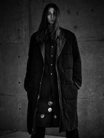 Monochrome portrait of a long-haired adult male wearing a long button-up coat and dress pants in front of a textured concrete wall.