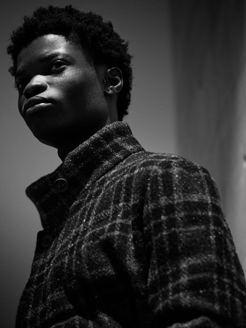 Black and white portrait of a young adult Black male in a patterned coat looking up and to the left against a plain background.