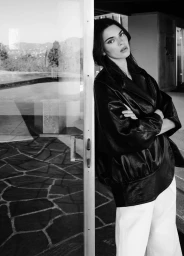 Kendall Jenner leaning against a glass door wearing a dark leather jacket and light trousers in a black and white campaign image.