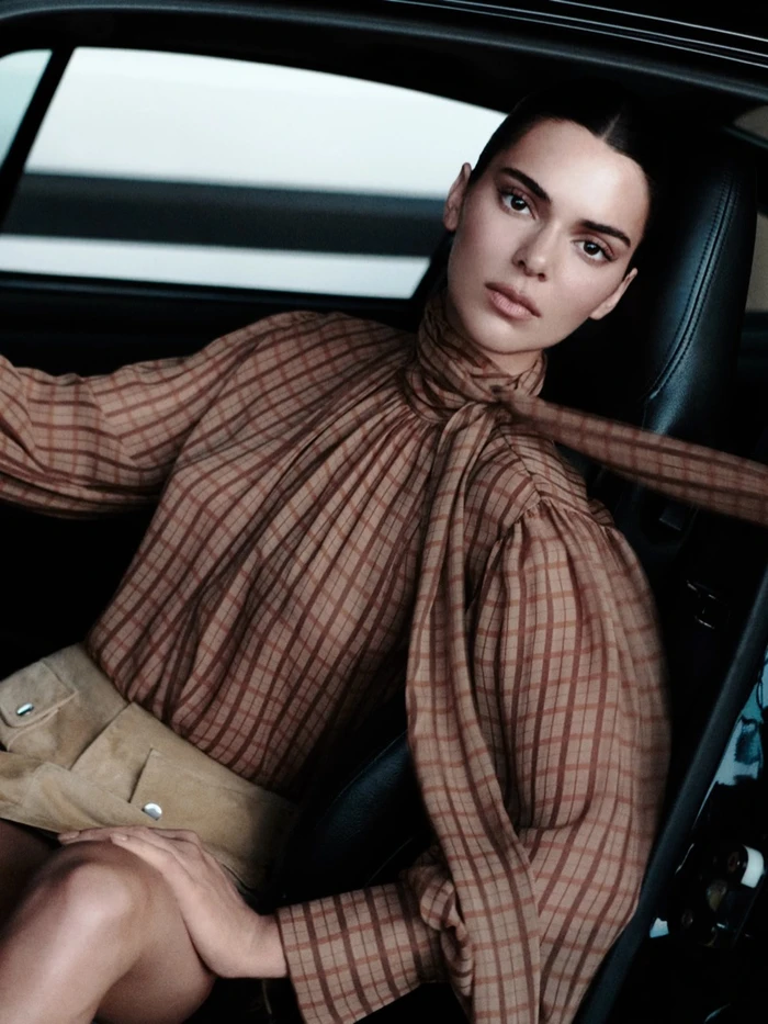Kendall Jenner sitting inside a car wearing a brown plaid blouse and tan skirt for the FRAME Fall 2026 campaign.