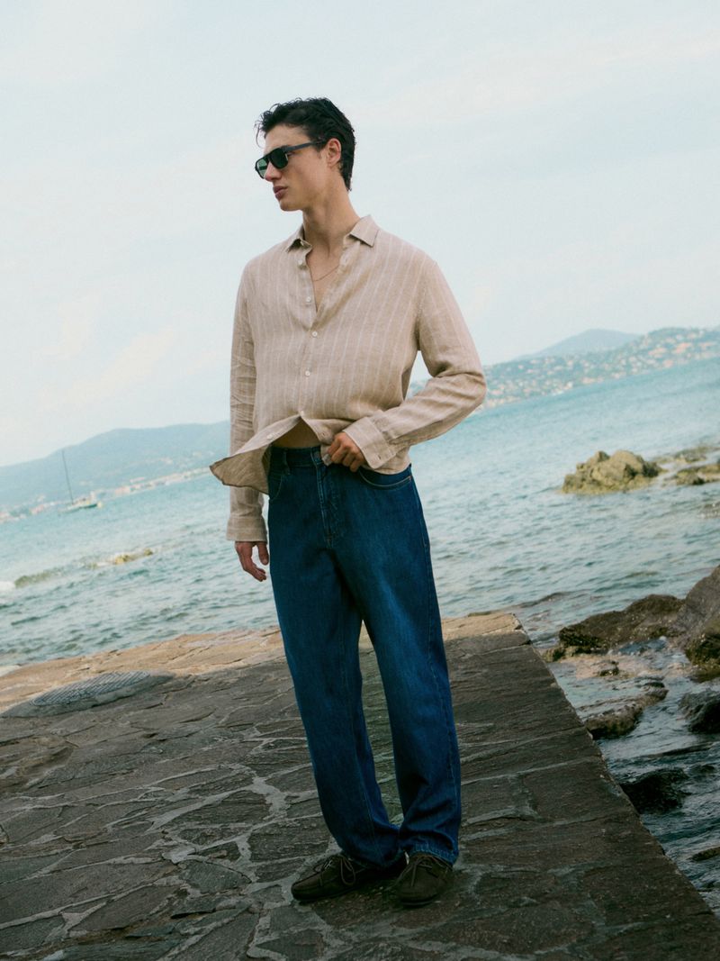 Adult male wearing jeans and an unbuttoned beige shirt standing on a stone structure with water and mountains in the background.