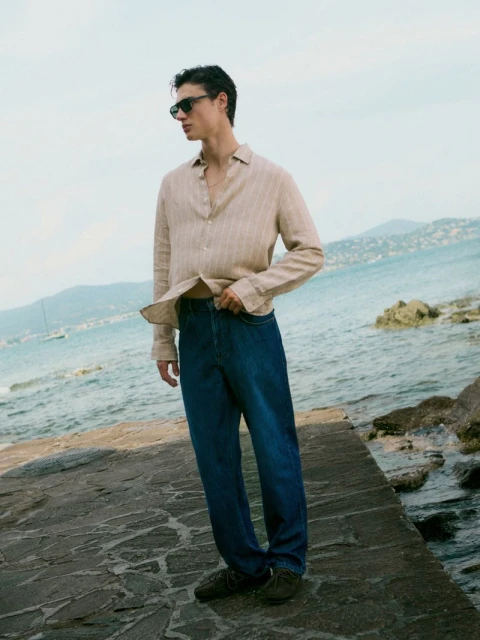 Adult male wearing jeans and an unbuttoned beige shirt standing on a stone structure with water and mountains in the background.