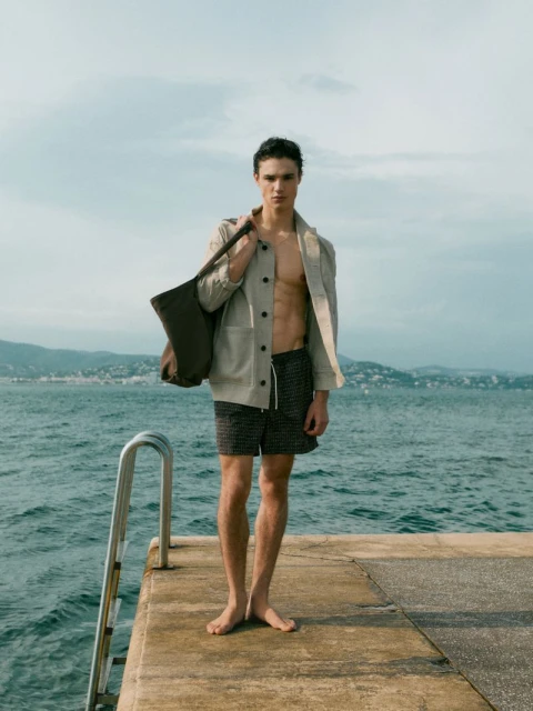 Athletic man in jacket and shorts stands on a dock overlooking the sea, carrying a tote bag with a clear sky and distant coastline.
