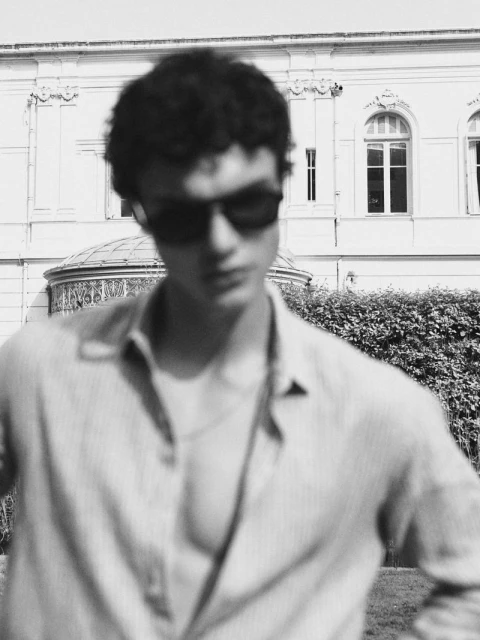 Black and white portrait of a young adult male with sunglasses, wearing an unbuttoned shirt outdoors with a building in the background.