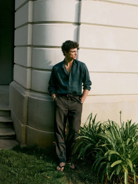 A young adult male poses near a building in a casual dark green button-down shirt and dark pants in an outdoor setting.