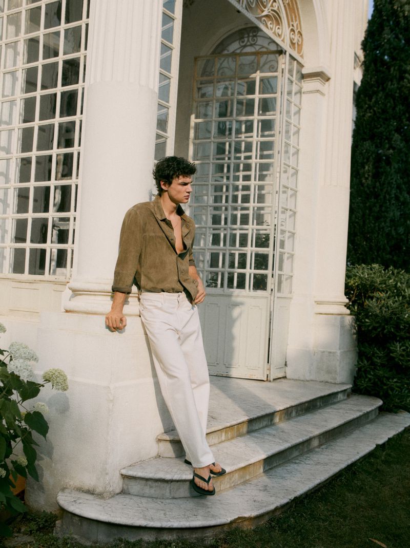 Man in olive shirt and white pants leans against a white column of a white building on a sunny day with greenery in the background.