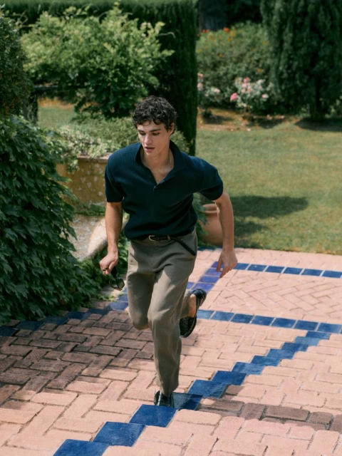 Young adult man in a polo shirt and trousers runs up brick stairs with blue tile accents, surrounded by green foliage in daylight.