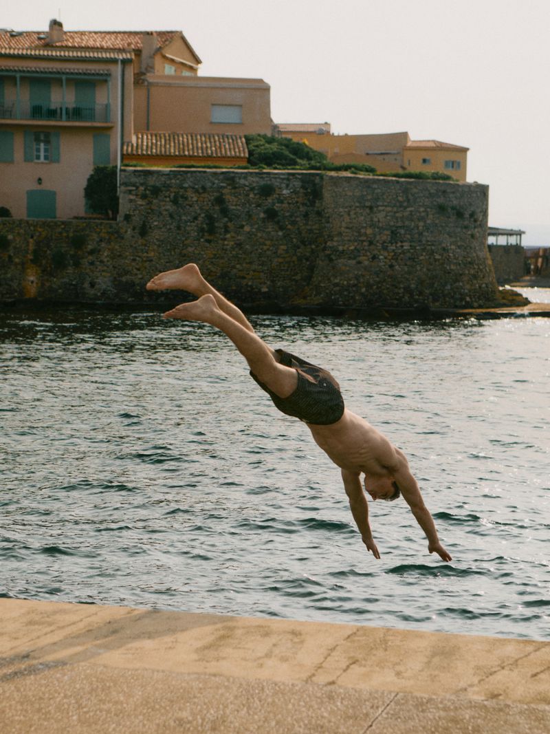 Man dives into ocean water near a stone wall and buildings. The sea is turquoise, and the sky is light blue on a sunny day.