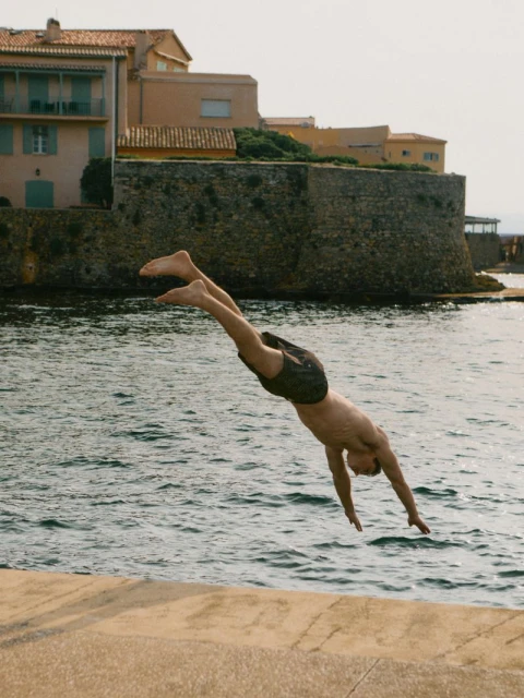 Man dives into ocean water near a stone wall and buildings. The sea is turquoise, and the sky is light blue on a sunny day.