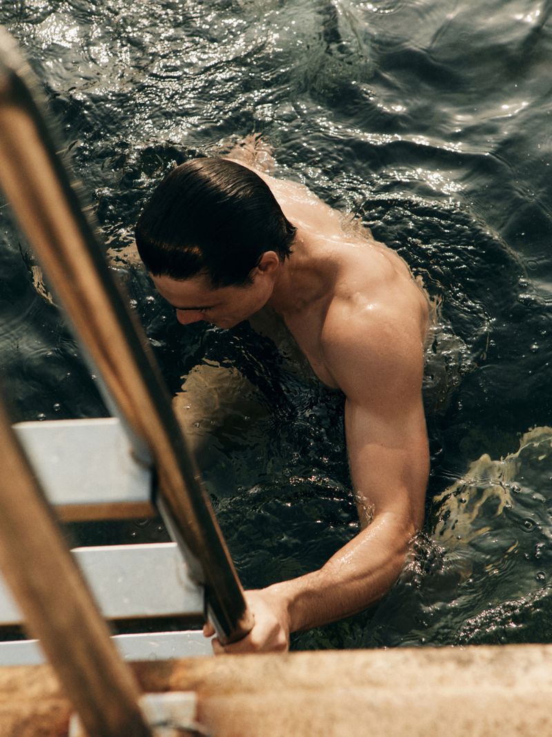 Athletic man climbs out of rippled water using ladder; warm tones, medium shot from high angle highlighting strength and definition.