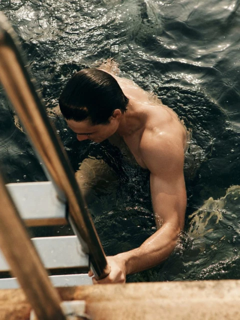 Athletic man climbs out of rippled water using ladder; warm tones, medium shot from high angle highlighting strength and definition.
