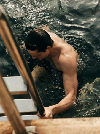 Athletic man climbs out of rippled water using ladder; warm tones, medium shot from high angle highlighting strength and definition.
