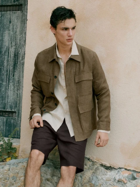 An adult male fashion model poses in casualwear, including shorts and an open jacket, leaning against a textured wall in a natural outdoor setting.