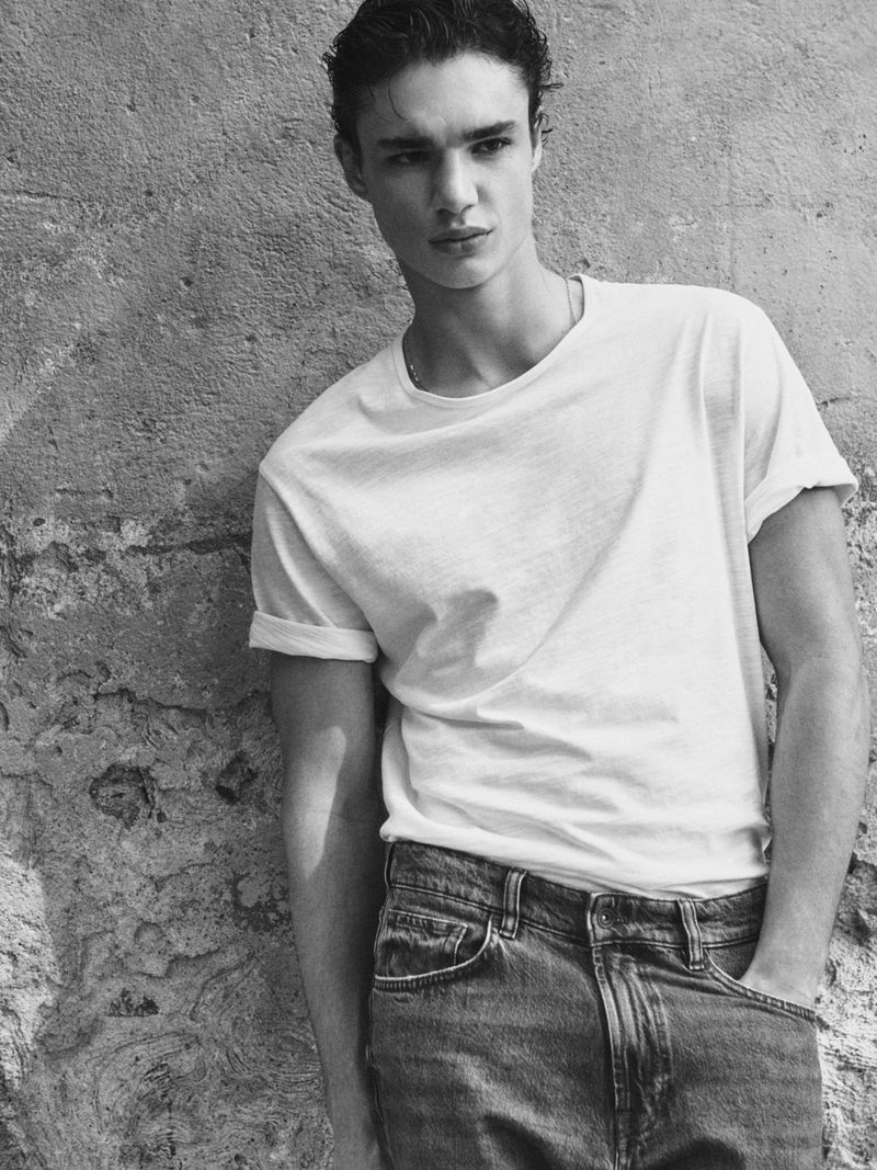Monochrome portrait of a young adult male model in a white t-shirt and jeans posing outdoors against a textured stone wall.