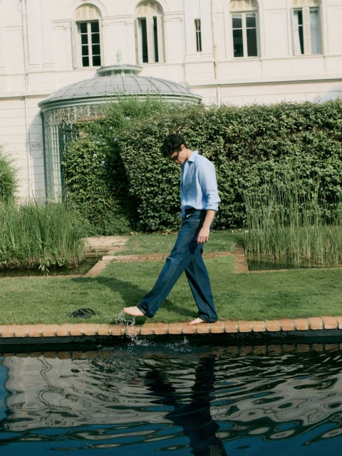 Young man in blue shirt walks barefoot on pool's edge, creating a splash. Building and greenery in the background, daylight scene.
