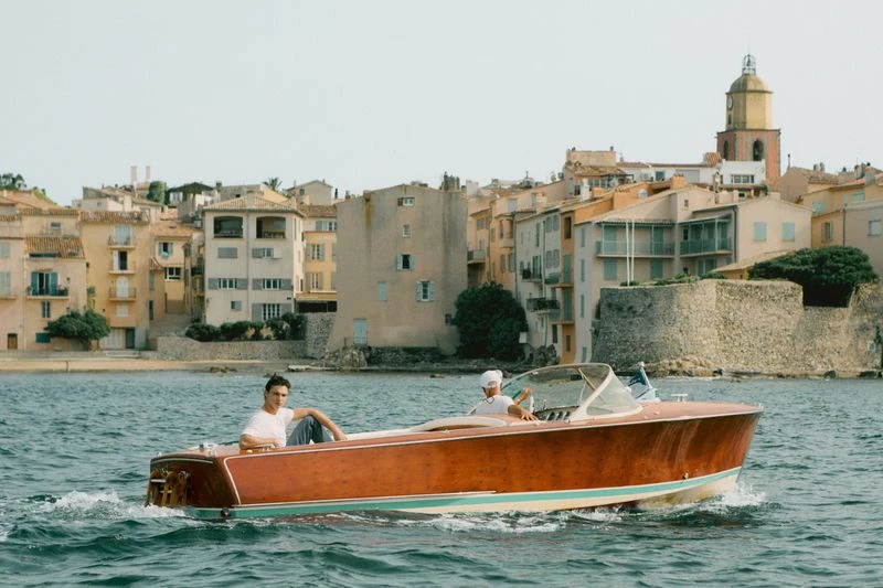 Two men on a wooden speedboat cruising on the water with a coastal town featuring houses and a clock tower in the background on a sunny day.