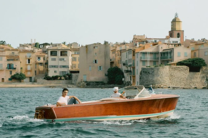 Two men on a wooden speedboat cruising on the water with a coastal town featuring houses and a clock tower in the background on a sunny day.