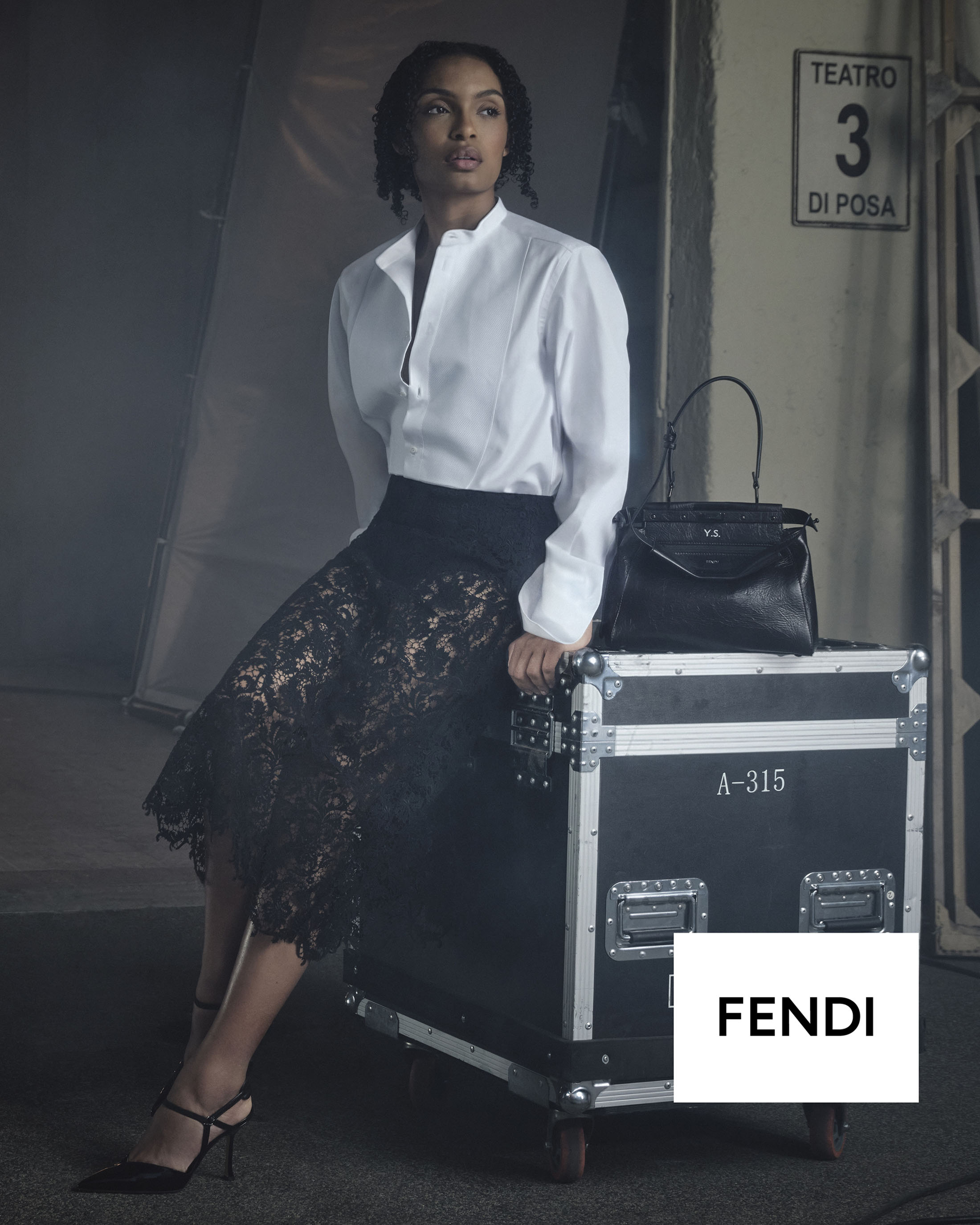 Yara Shahidi seated on a black flight case wearing a white shirt and black lace skirt beside a Fendi Peekaboo bag in a studio.