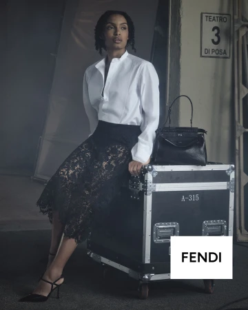 Yara Shahidi seated on a black flight case wearing a white shirt and black lace skirt beside a Fendi Peekaboo bag in a studio.