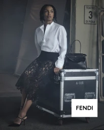 Yara Shahidi seated on a black flight case wearing a white shirt and black lace skirt beside a Fendi Peekaboo bag in a studio.