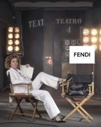Valeria Golino seated in a director's chair in a white pantsuit with a black Fendi Peekaboo handbag on a film set.
