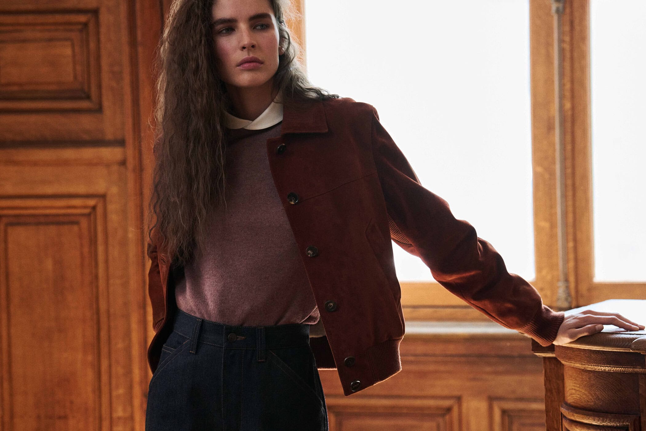 Model wearing a rust brown suede button-up jacket, knit sweater, and dark trousers indoors beside wooden panelling and a window.