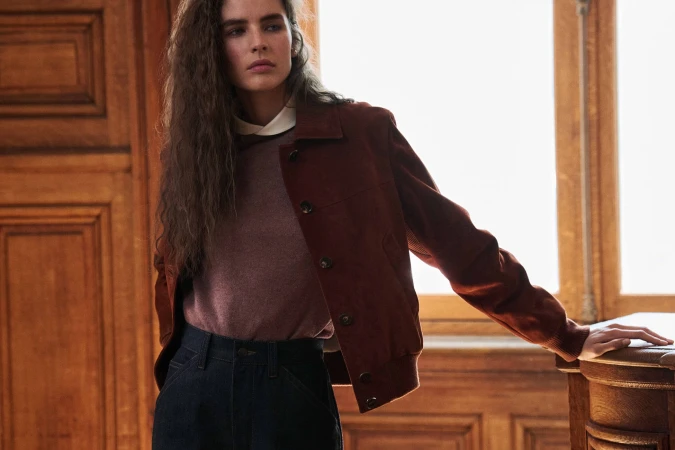 Model wearing a rust brown suede button-up jacket, knit sweater, and dark trousers indoors beside wooden panelling and a window.