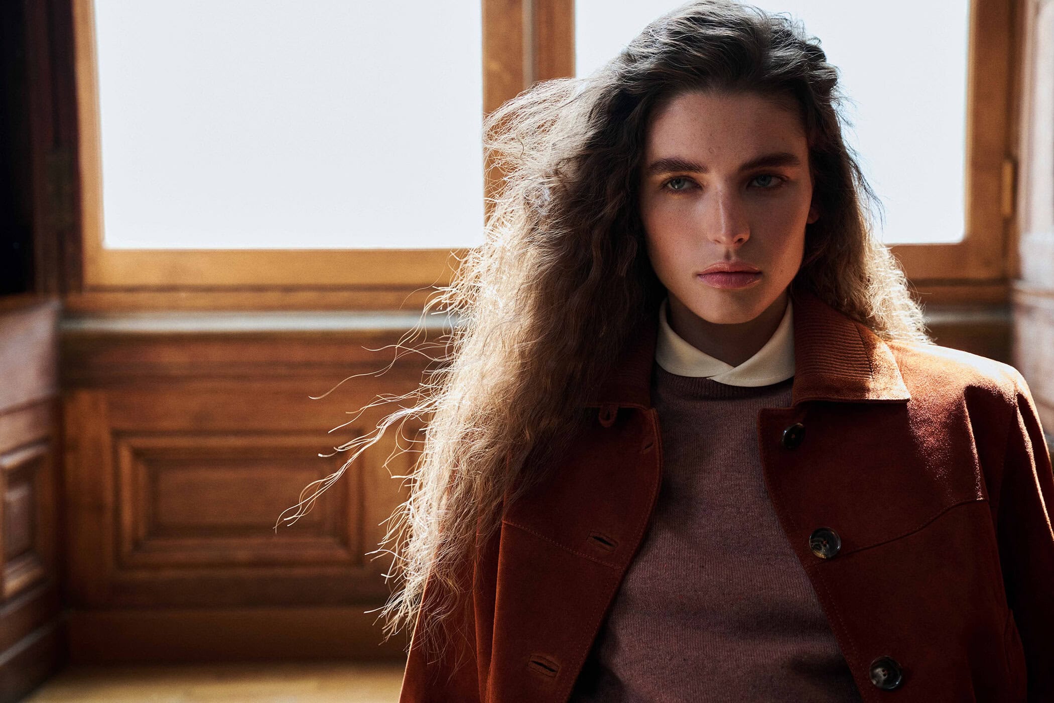 Model Libby Taverner wearing a rust suede jacket over a knit sweater in front of wood wall paneling and a large window.