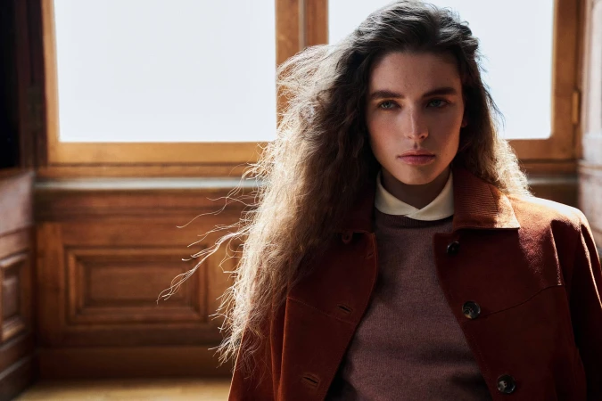 Model Libby Taverner wearing a rust suede jacket over a knit sweater in front of wood wall paneling and a large window.