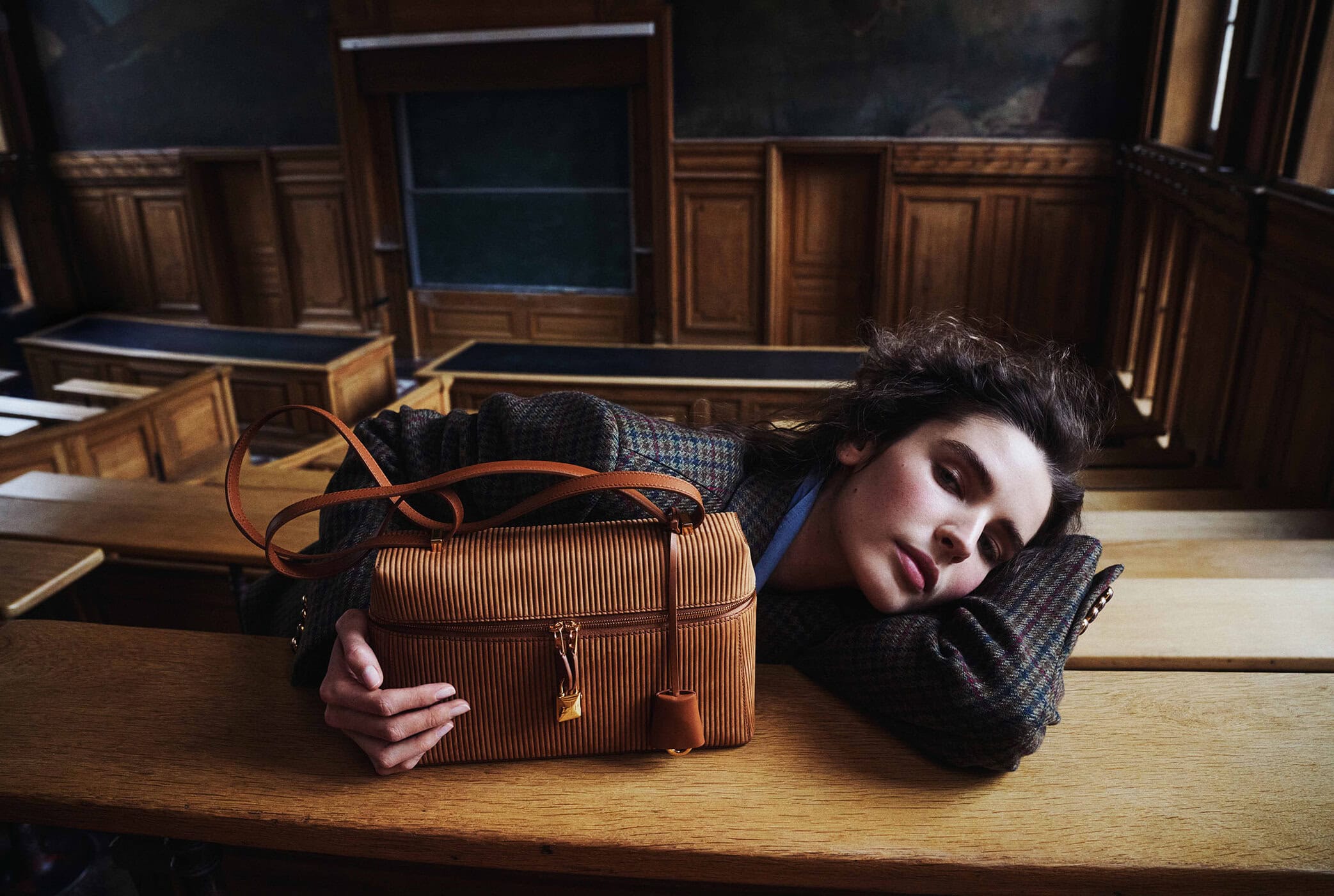 Model Libby Taverner resting her head on a wooden desk next to a ribbed camel leather handbag in a wood-paneled lecture hall.