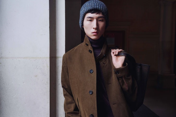 Kim Woosang wearing a brown wool coat, dark turtleneck, and grey knit beanie, holding a shoulder bag in a stone doorway.