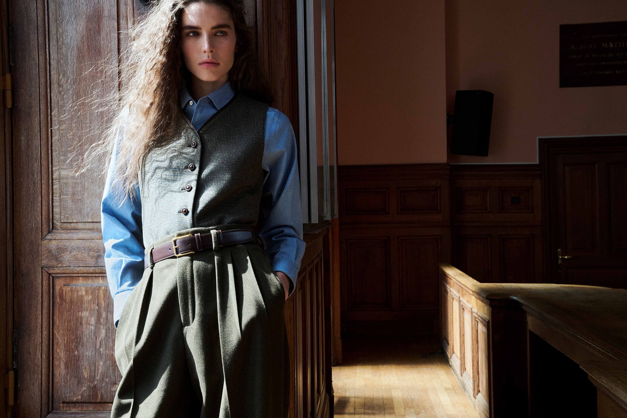 Model Libby Taverner wearing an olive vest, blue shirt, and pleated trousers in a wood-paneled room for Loro Piana.