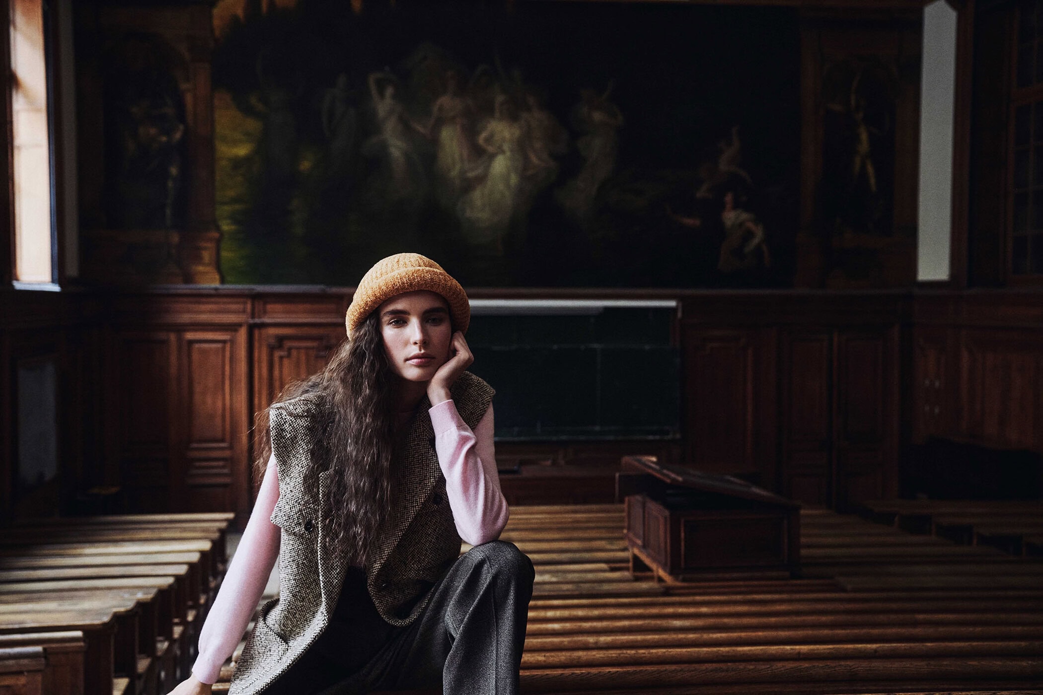 Woman in an orange knit beanie and tweed vest seated on wooden lecture hall benches at the Sorbonne in Paris.
