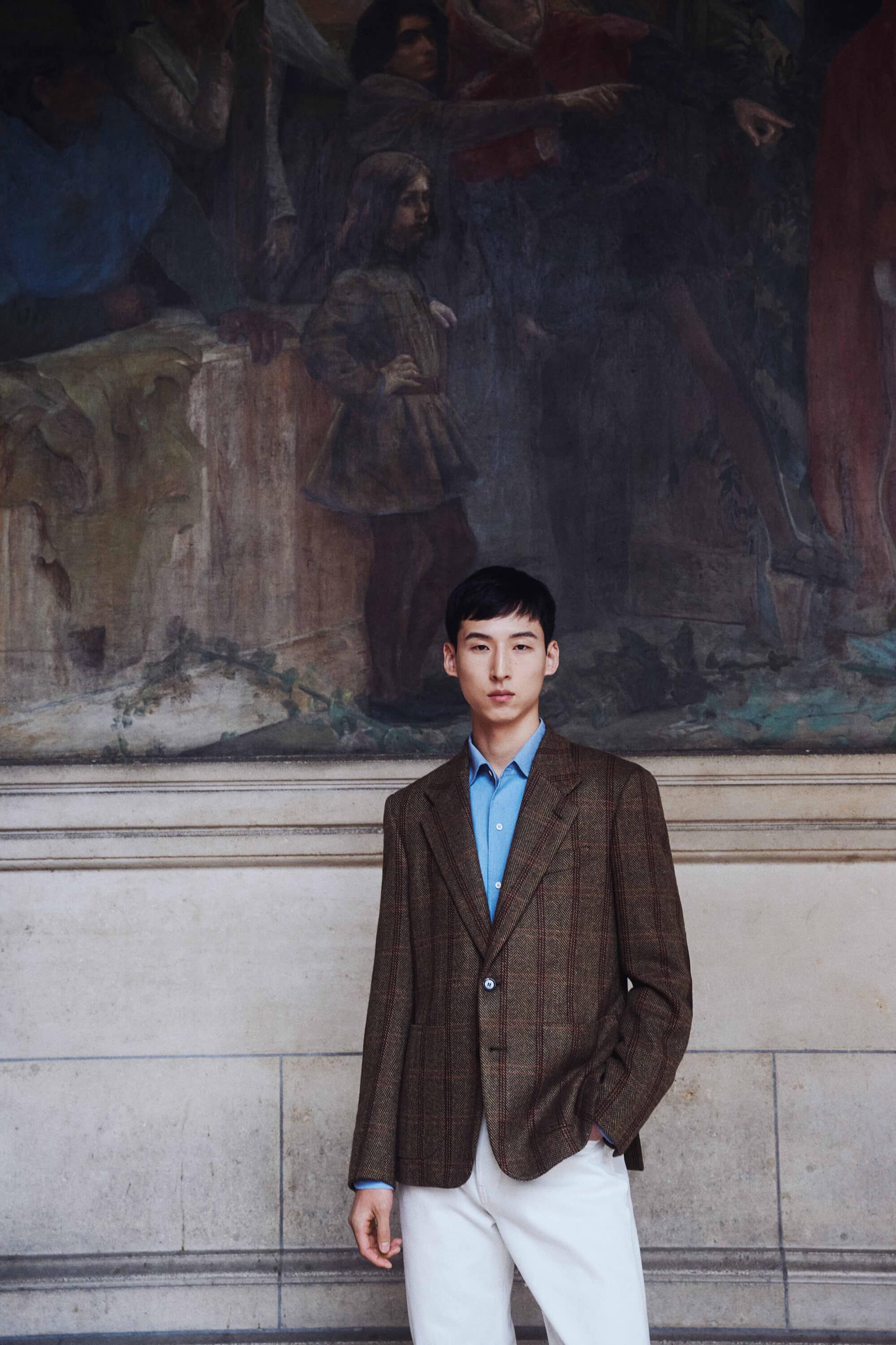 Kim Woosang wearing a brown patterned blazer, blue shirt, and white trousers in front of a historic painted mural at Sorbonne, Paris.